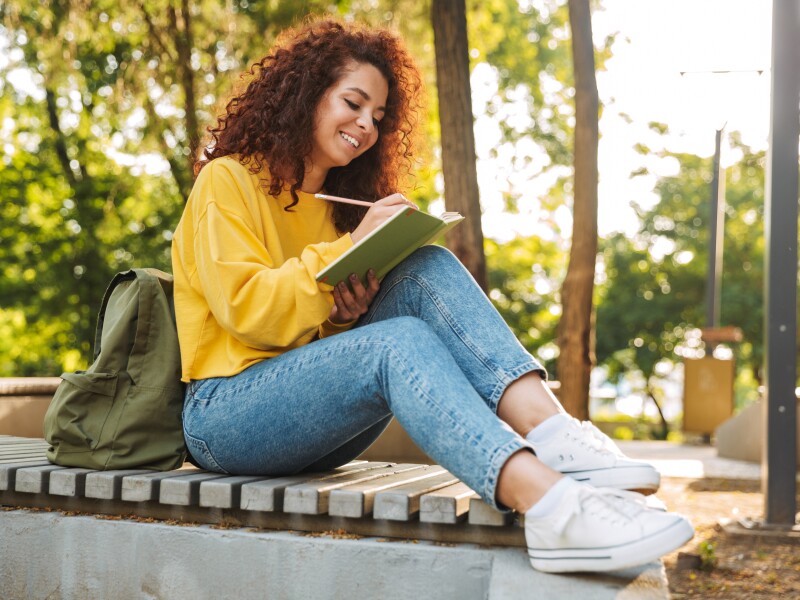 Young beautiful curly student girl sitting outdoors in nature park writing notes in notebook.