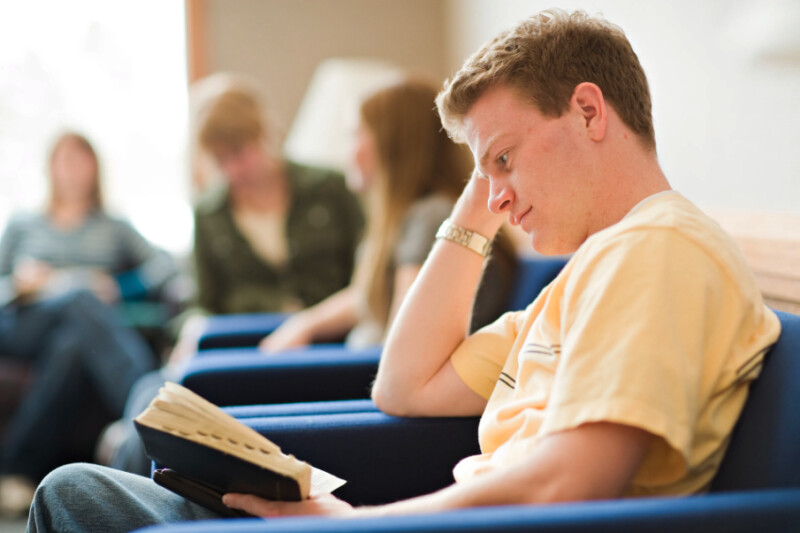 A young man sits in a chair and reads from his scriptures.