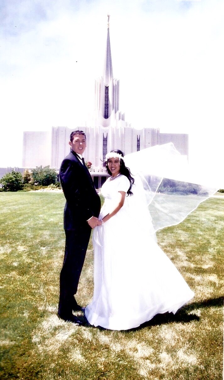 A young man and a young woman wearing wedding clothes smile in front of the Jordan River Utah Temple.
