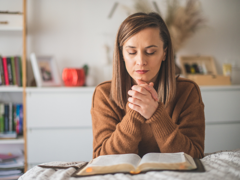 A woman prays next to her bed near her open scriptures