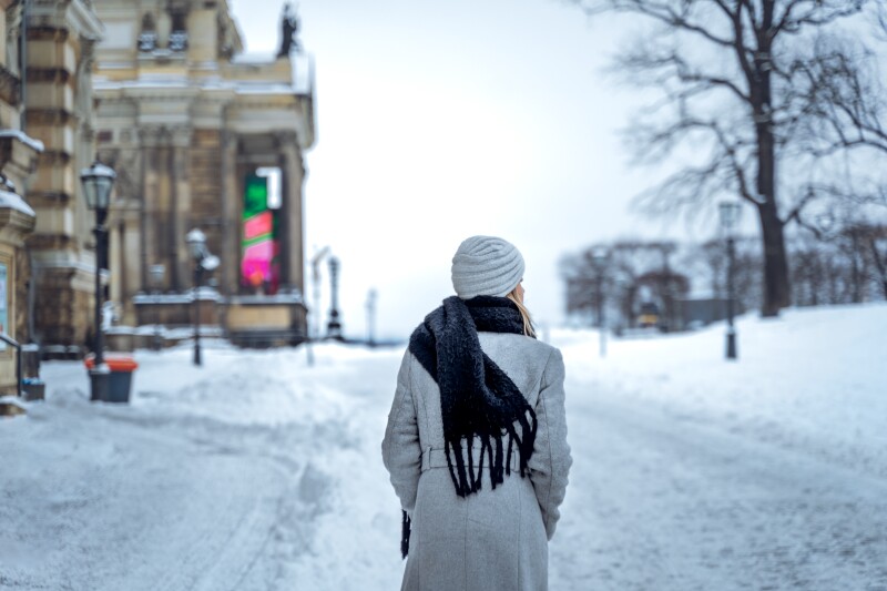 Back view of anonymous female in coat with scarf and hat holding hands in pockets and looking away while strolling on snowy road on winter day in Dresden, Germany.