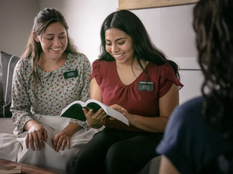 Two sister missionaries sitting on a couch smile while looking at a scripture verse.