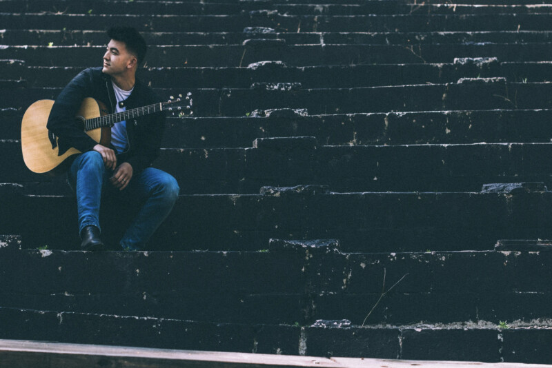 Brandon Pak holds a guitar sitting on stair steps.