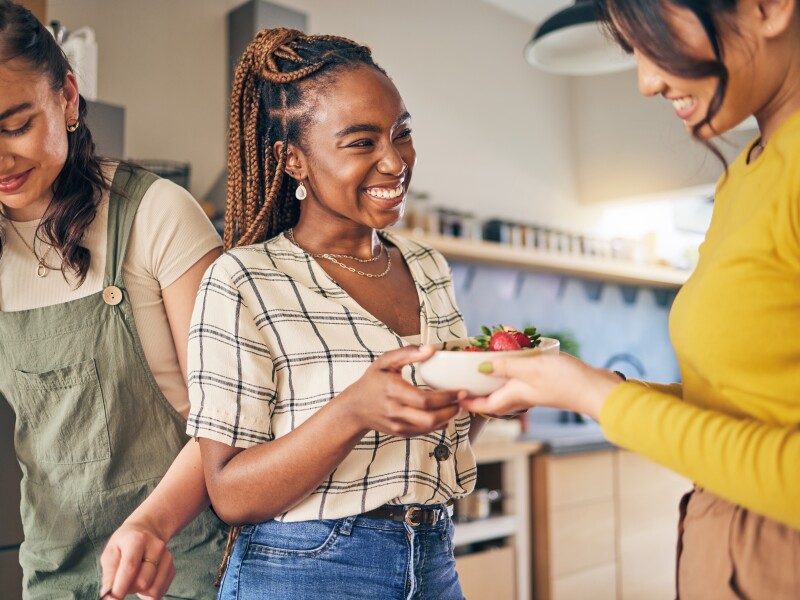Women, friends and eating fruit in home for bonding, nutrition and happy lunch together. Healthy diet, sharing and wellness, fresh summer food for friendship and girls in kitchen with smile at party.