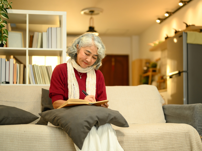 Calm senior woman in a red sweater sitting on couch and writing in journal