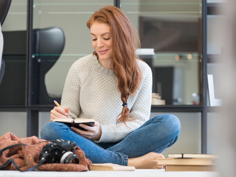 A woman with red hair sits on the floor and smiles while writing in a journal.