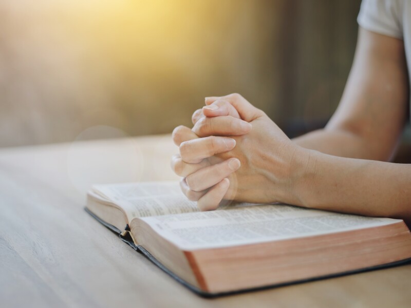 Close up of a woman hands praying on an open bible on a wooden table with window light Bokeh.