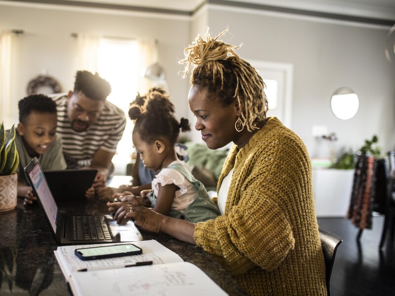 Mother working from home while holding toddler, family in background