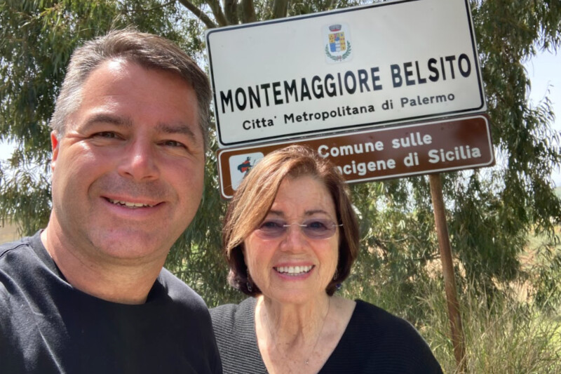 Grant and Geri Guinn smile in front of a sign for Montemaggiore Belsito near Palermo, Sicily.