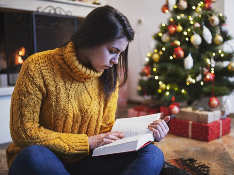 Young Beautiful Woman is Reading a Book Next to the Christmas tree and the Fireplace at Home