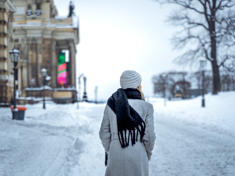 Back view of anonymous female in coat with scarf and hat holding hands in pockets and looking away while strolling on snowy road on winter day in Dresden, Germany.