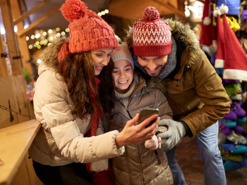A mother, daughter, and father wearing winter clothes at a Christmas market smile looking at a mobile phone together.