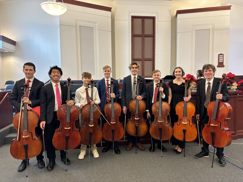 Eight youth holding cellos smile in a Latter-day Saint chapel at Christmastime.