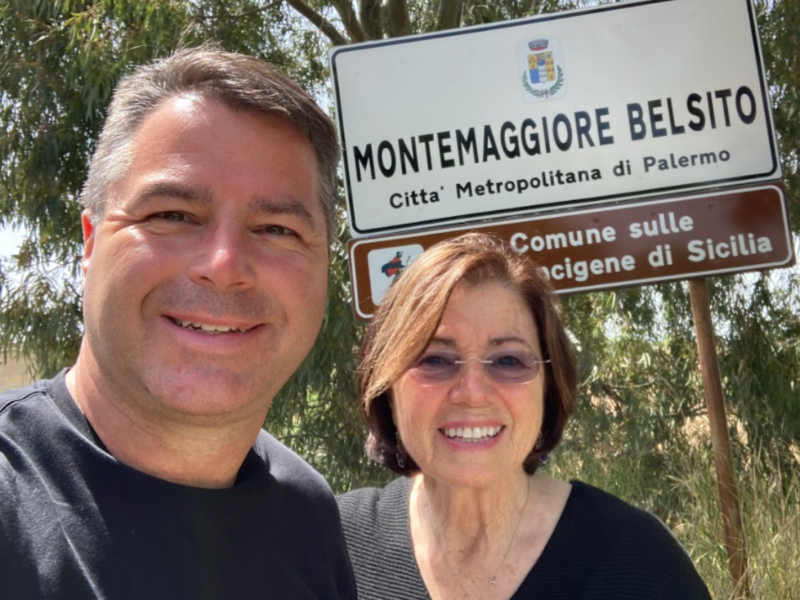 Grant and Geri Guinn smile in front of a sign for Montemaggiore Belsito near Palermo, Sicily.
