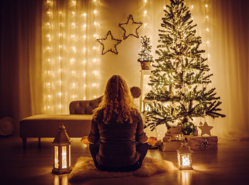 A woman sitting on a sheepskin rug meditates in front of a Christmas tree.