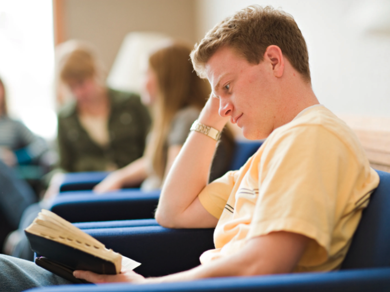 A young man sits in a chair and reads from his scriptures.