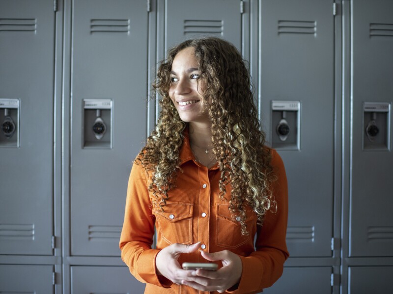 Female high school student standing near lockers holding phone