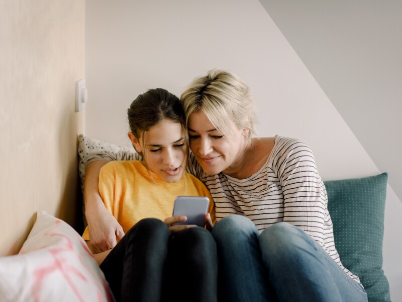 A mother and a daughter smile together looking at a phone screen.