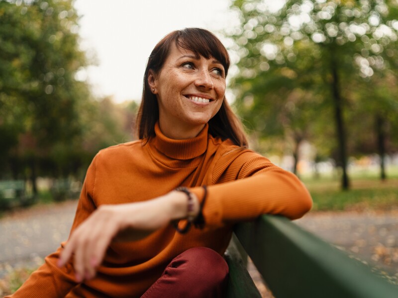 Beautiful mid adult woman sitting on bench and resting outside in the city on autumn day.