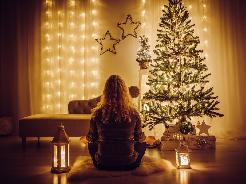 A woman sitting on a sheepskin rug meditates in front of a Christmas tree.