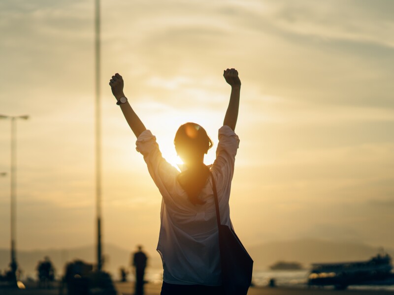 Young woman relaxing with hands in the air by the pier and enjoying the beautiful sunset and warmth of sunlight