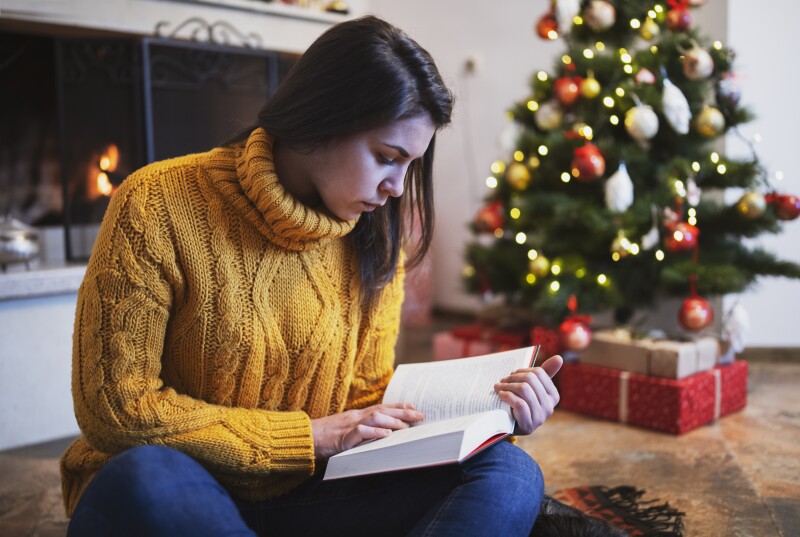 Young Beautiful Woman is Reading a Book Next to the Christmas tree and the Fireplace at Home