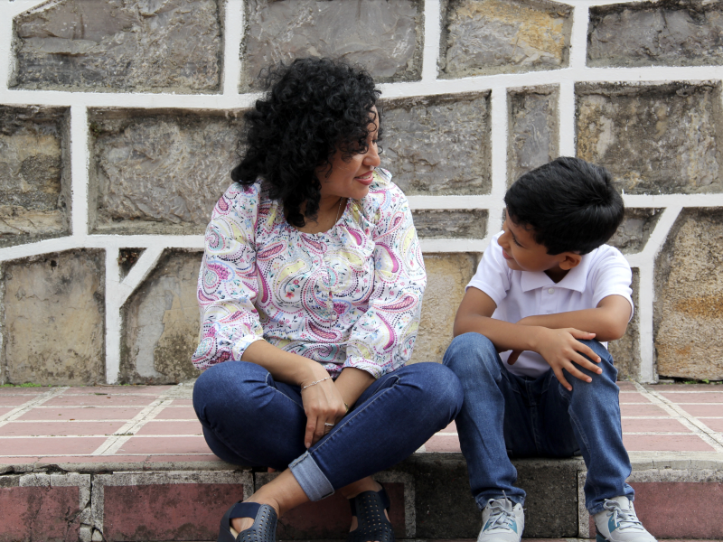 Dark-haired Latin mom and son smile at each other while sitting outside on a step.