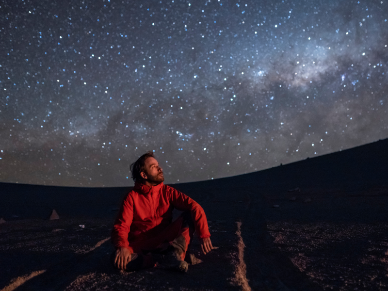 A young adult man seating and looking at the view of our Milky Way galactic core located in the constellation of Sagittarius, an amazing view at Atacama Desert.