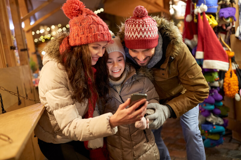 A mother, daughter, and father wearing winter clothes at a Christmas market smile looking at a mobile phone together.