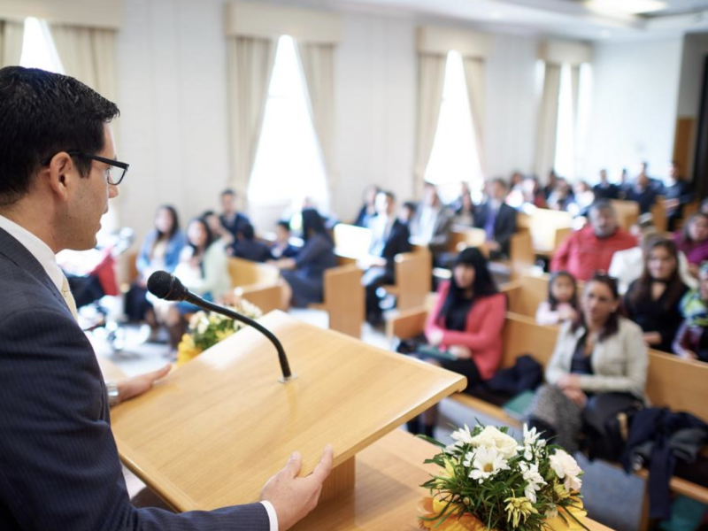 A man stands at the pulpit in a Latter-day Saint chapel and speaks to the congregation.