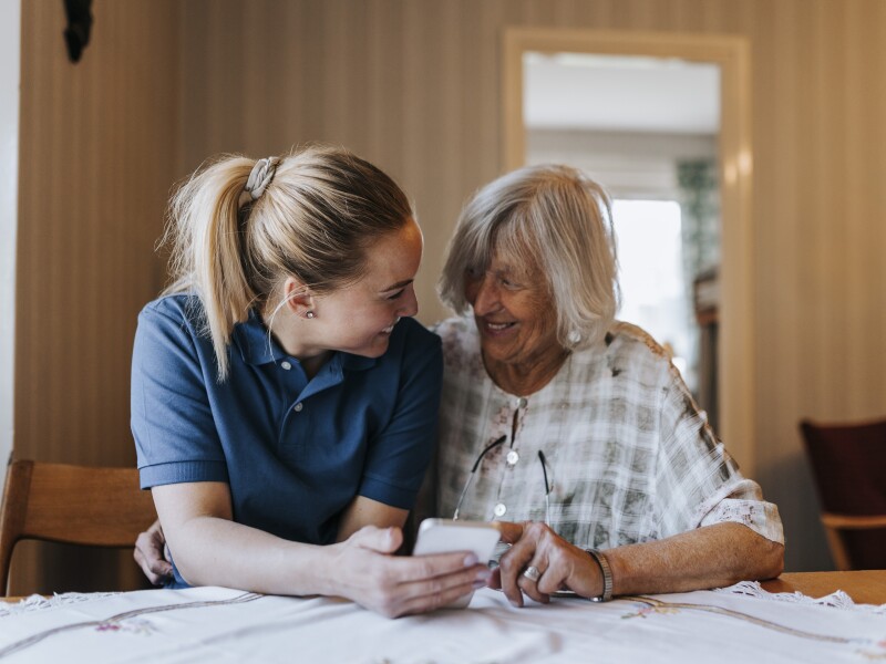 Smiling female care assistant sharing smart phone with senior woman while sitting at dining table in home
