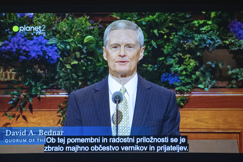 A screenshot of the April 6, 2025, PlanetTV broadcast in Slovenia of Elder David A. Bednar speaking in April 2025 general conference. The transcription of his message—in Slovenian—is “A small congregation of believers and friends gathered for this momentous and joyous occasion.”