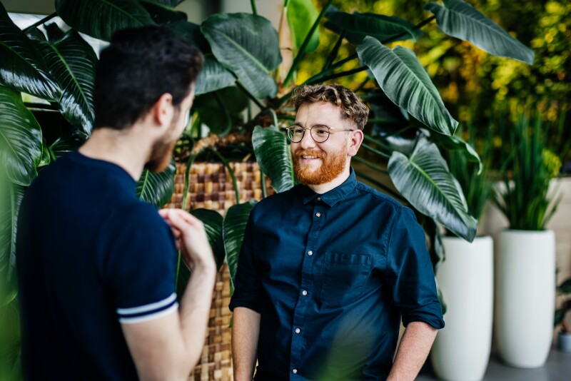 Two Colleagues Standing Amongst Potted Plants In Green Office Space, Talking