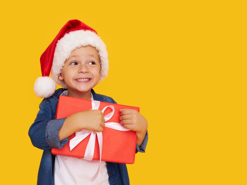 Portrait of a satisfied little child boy in christmas Santa hat. laughing isolated over yellow background. Holds a gift box. Preparing for the New Year holidays