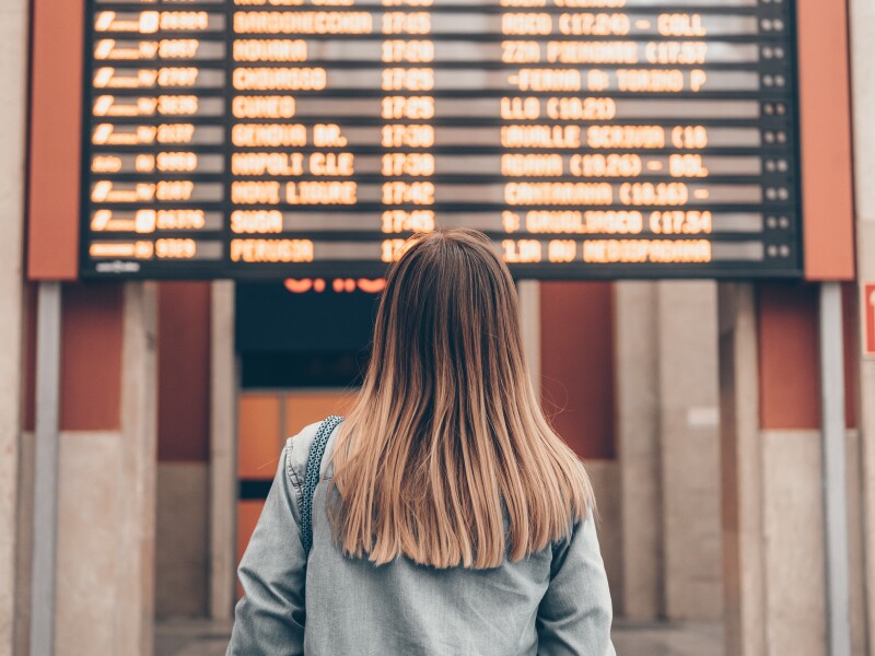 A young woman at a railway station or at the airport looks at the smartphone screen against the background of the arrival and departure board