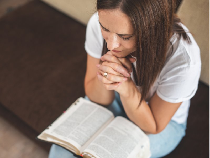 A young woman prays with an open Bible on her lap.